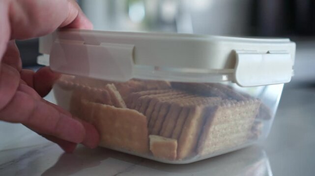 hand placing a plastic container filled with crackers on a table. Kitchen footage showing food storage, snacks, and everyday home lifestyle.