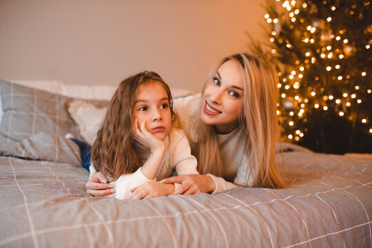 Happy woman with child girl in bed over Christmas tree with lights in room. Winter holiday season.