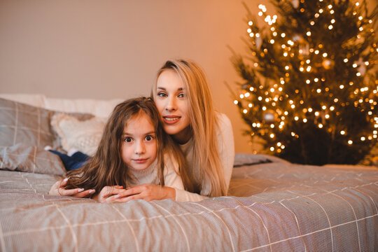 Happy woman with child girl in bed over Christmas tree with lights in room. Winter holiday season.
