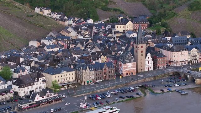 Slow aerial approach toward the old town of Bernkastel-Kues, Germany, featuring St. Michael&rsquo;s Tower, traditional riverside buildings and vineyard-covered hills above the Moselle Valley.