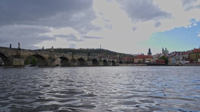 Wide scenic view of Charles Bridge crossing the Vltava River in Prague, with historic stone arches, riverside architecture and the city skyline beneath a dramatic cloudy sky