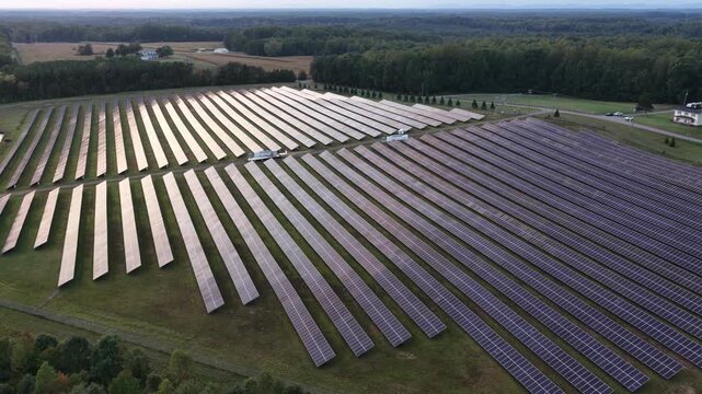 Parallel solar panels fill the foreground in Louisa County, Virginia, opening onto farmland, scattered homes and woodland beneath a pale evening sky. Aerial view