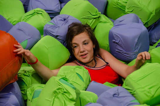 Smiling young girl sitting in foam cube ball pit. Teenager jumping onto obstacle in indoor adventure park. Teenager standing in indoor trampoline park. Young girl jumping into foam pit at trampoline