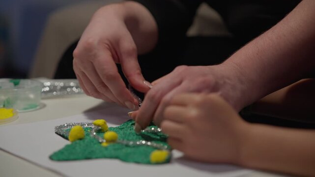 An adult's hands and a child's hands attach silver tinsel and yellow pom poms to a glittery green star-shaped Christmas craft on a table