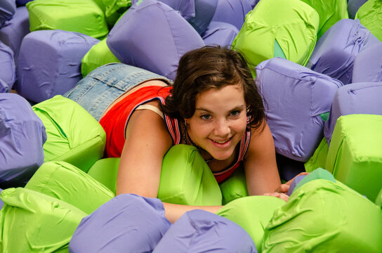 Smiling young girl sitting in foam cube ball pit. Teenager jumping onto obstacle in indoor adventure park. Teenager standing in indoor trampoline park. Young girl jumping into foam pit at trampoline