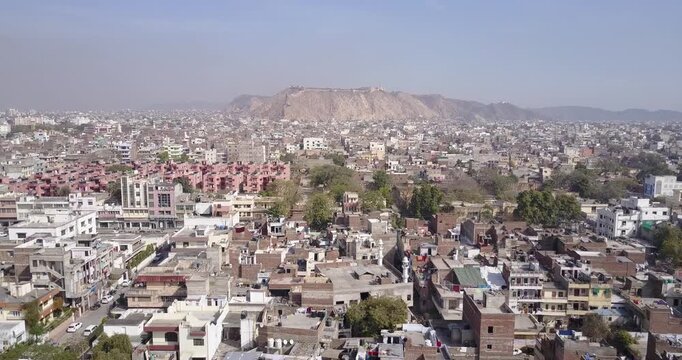 Dense Jaipur neighborhood of flat roofs and pink apartment blocks with the Nahargarh Fort ridge on the hazy horizon as the camera drifts sideways, Aerial View