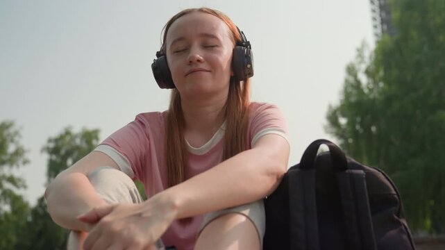 Young woman sits outdoors wearing headphones and pink shirt with her arms resting on bent knees. Woman appears relaxed in soft daylight against urban park backdrop with trees and distant
