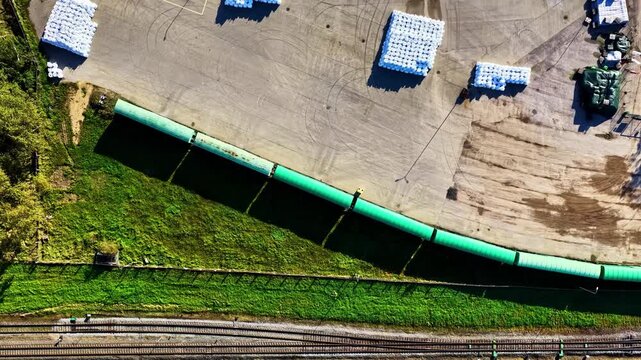Aerial view of a long green pipeline running parallel to railway tracks through a rural landscape in Eleja, Jelgava Municipality, Latvia.