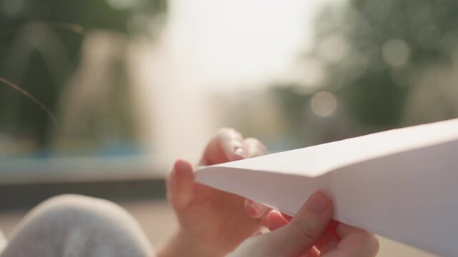 Young woman carefully folds white paper airplane adjusting its shape for optimal flight. soft natural light creates gentle shadows on her hands and smooth texture of folded material
