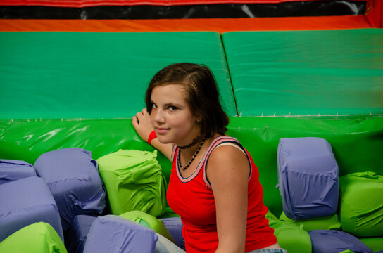 Smiling young girl sitting in foam cube ball pit. Teenager jumping onto obstacle in indoor adventure park. Teenager standing in indoor trampoline park. Young girl jumping into foam pit at trampoline