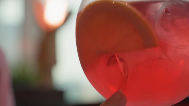 Close up of hand gently touching side of red cocktail glass with orange slice emphasizing texture and detail. liquid inside is vibrant pinkish red in color creating visual interest