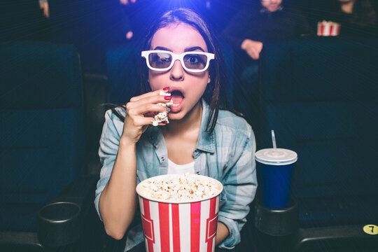Amazed girl is sitting very close and looking straight. She wears special 3d glasses for watching movies. Girl is eating popcorn with her right hand. Also there is a cup of coke besides her.