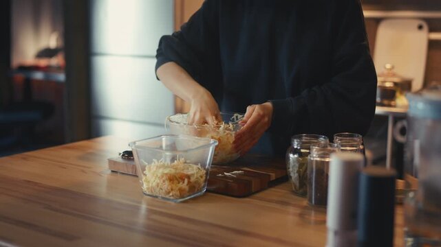 Woman hands putting seasoned shredded cabbage and carrots from a bowl into a glass container. Preparing homemade fermented sauerkraut on a wooden table in a cozy warm home kitchen. Close-up process.