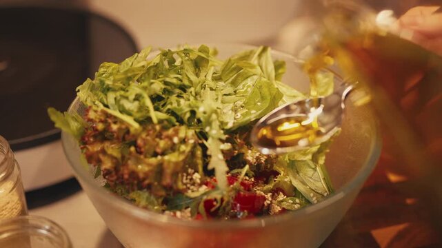 Close-up of a person pouring olive oil from a glass oil jug into a spoon to dress a fresh salad with lettuce, arugula, chopped tomatoes, basil, and sesame seeds in a glass bowl in a warm home kitchen.