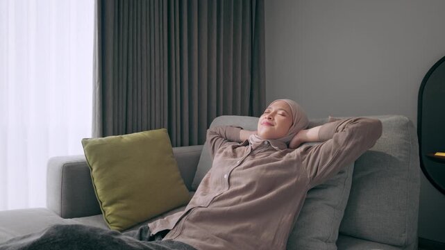 Asian Muslim woman wearing a hijab leans back on a comfortable sofa in a modern living room, eyes closed and smiling as she enjoys a quiet moment of relaxation and calm at home.