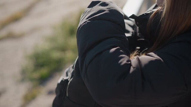 Woman with long brown hair holds black backpack adjusting zipper while wearing dark jacket. background is softly blurred outdoor scenery with natural light and muted colors emphasizing her