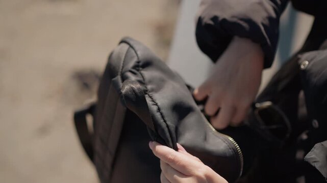 Woman carefully organizes her belongings in black bag handling items like sunglasses and zippers. Woman wears dark outerwear with visible texture details such as stitching or fabric weave