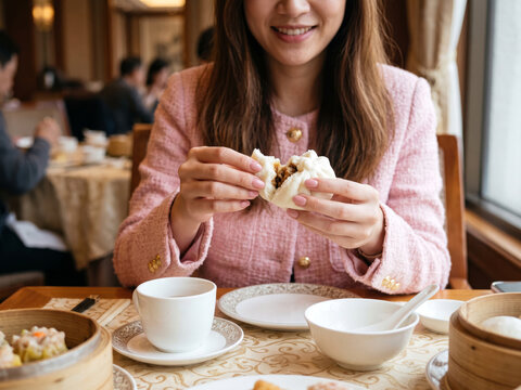Asian woman opening freshly steamed pork bao bun enjoying dim sum breakfast at Chinese restaurant.