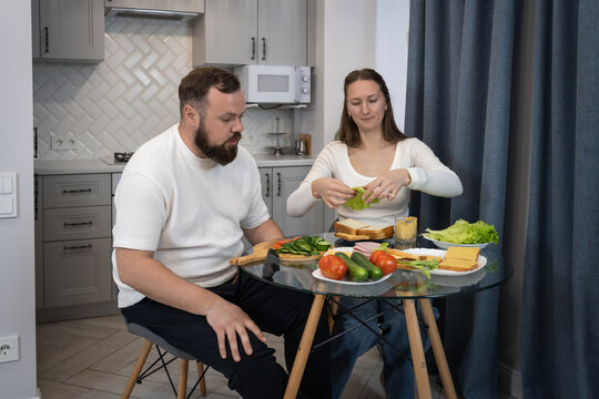 Young couple preparing a healthy meal together, slicing fresh vegetables and assembling sandwiches at the kitchen table, sharing a moment of domestic life