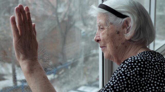 Lonely senior woman with gray hair looking out the window during quarantine, putting her hand on glass to say goodbye to her family, symbolizing social distancing, isolation, and nostalgia for past