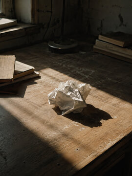 A crumpled ball of white paper lies on a weathered wooden table in a dimly lit room, hinting at a moment of frustration.