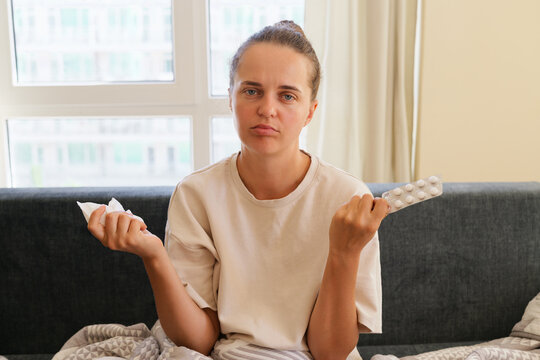 Sick frustrated young woman sitting on sofa wrapped in blanket holding tissue napkin and blister of pills gesturing with hands during seasonal flu illness at home