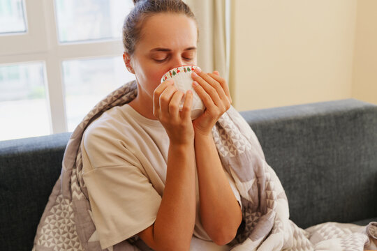 Close up portrait of sick young woman wrapped in blanket holding mug with both hands and drinking hot tea to treat throat pain during seasonal flu at home