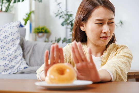 glazed donuts on plate with Asian woman making stop hand gesture in background, refusing unhealthy sugary food for diet and weight loss healthy concept.