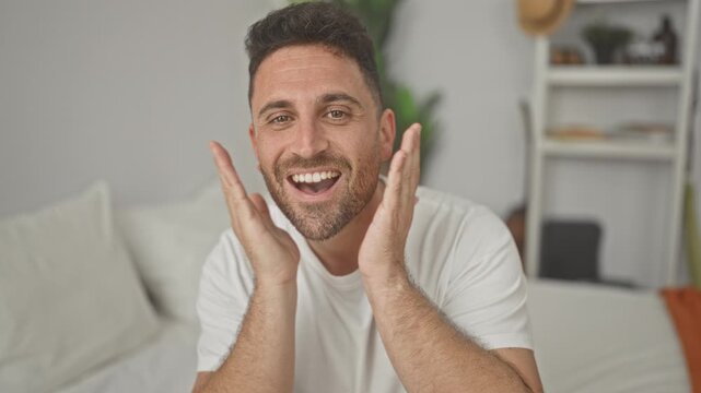 Man on bed smiling and touching his face with one hand on forehead and palms to cheeks in a bedroom; joy.