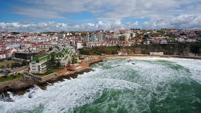Majestic aerial view of the charming coastal town of ericeira in portugal, with large atlantic ocean waves crashing on the sandy shores of baleia beach on a beautiful, partly cloudy day