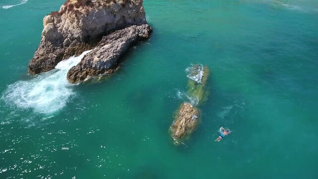 Aerial view of a man swimming in the crystal clear turquoise sea near scenic rock stacks on a sunny summer day in algarve, portugal, highlighting a refreshing and adventurous coastal getaway