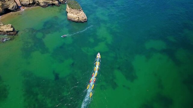 Dramatic top down drone shot of a group of colorful kayaks being towed by a boat across the emerald green ocean, alongside the stunning cliffs and rock formations of the algarve coast