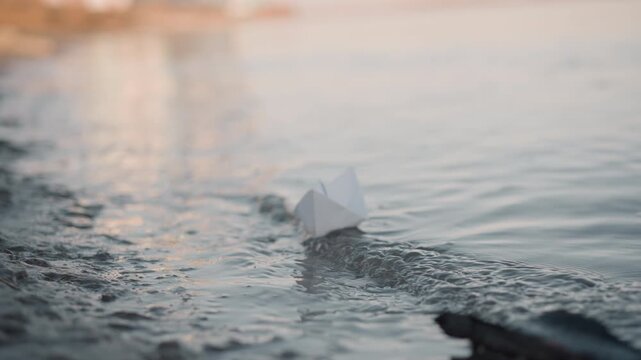 White paper boat gently drifts on calm water gradually moving away from shore. soft lighting creates peaceful atmosphere with subtle reflections and natural ripples in background. scene