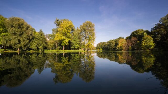 Time lapse of autumn foliage reflecting on calm lake water surface under sunny clear sky in city park