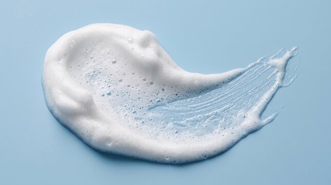 Elegant curved swoosh of thick white cleansing foam with tiny bubbles smeared across a smooth pastel blue background in macro studio light