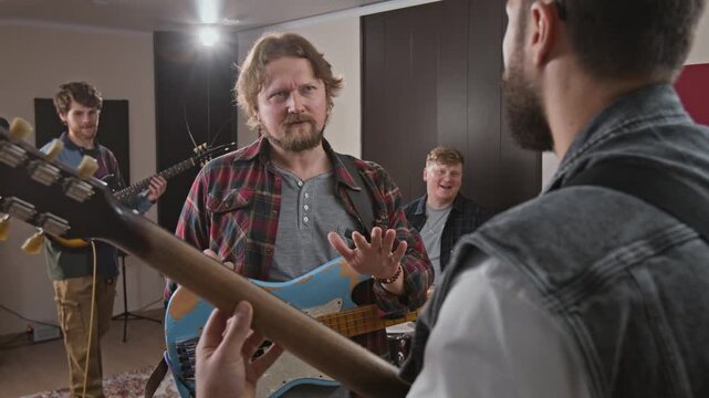 Caucasian young adult bassist discussing song arrangement with bandmates during rehearsal in music studio. Visual suits music production and creative team collaboration ads