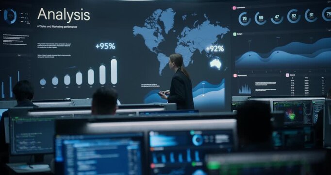 Wide Shot of Female Data Analyst With Tablet Studying Giant Sales Analysis Wall Screen in Modern Financial Control Room, While Analysts Monitor Global Trade, Banking Growth and Cybersecurity Feeds.