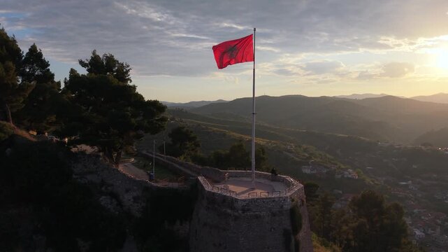 Aerial view of the Albanian flag waving atop the historic stone walls of Berat Castle during sunset in Berat, Berat County, Albania.