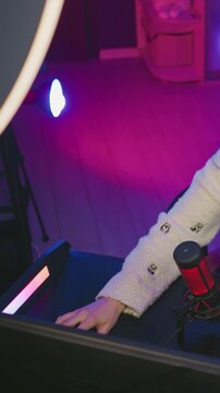 Young woman in white sweater with panda patterns interacts casually at her desk featuring vibrant pink and purple studio lighting. red microphone on table is visible as woman rests or moves