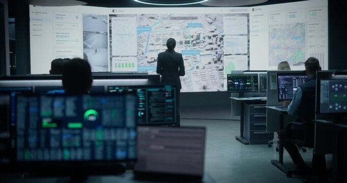 Logistics Company Monitoring Control Room People Work on Growth and Optimisation. Wide Shot of Female Operations Lead With Tablet Facing City Map Wall, Auditing Traffic Flow and Infrastructure Data