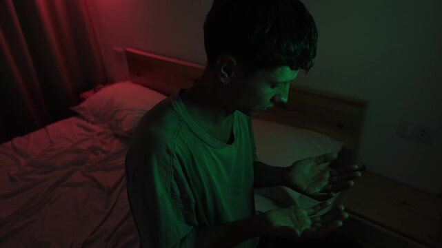 Dissociated man sitting alone on a bed examining his hands, turning his palms and clenching his fists while experiencing detachment from reality in red and green low key light