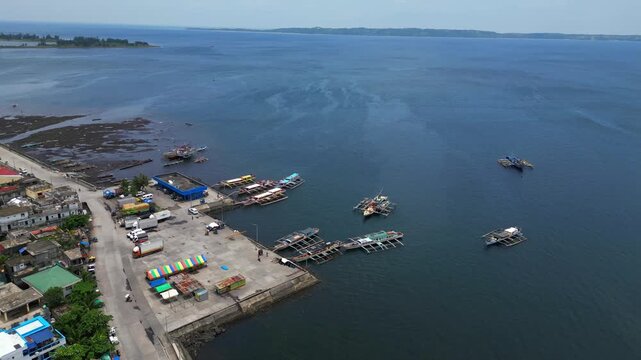 Aerial drone footage of San Roque Boulevard and the waterfront near Tabaco Port in Tabaco City, Albay, Philippines. View of the coastal road, port area, boats and urban shoreline under clear sunny ski