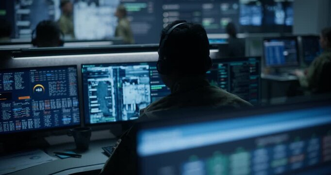 Army Military Surveillance Monitoring Control Room: Back View Portrait of Soldier in Headset Tracking Satellite Map Data on Multiple Displays, While Officers Hold Briefing at Big Screen in Background.
