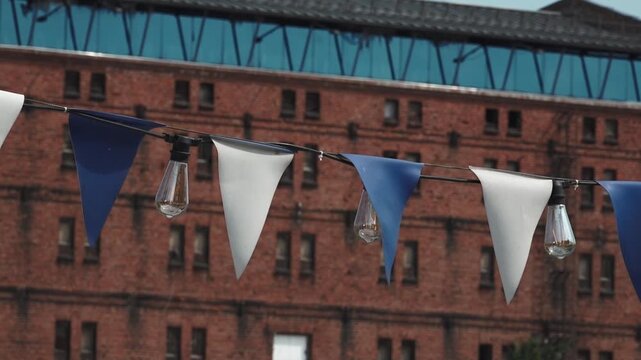 Close-up of blue and white triangular bunting flags fluttering in the wind beside hanging string light bulbs, with a red brick building and rows of windows softly blurred in the urban background