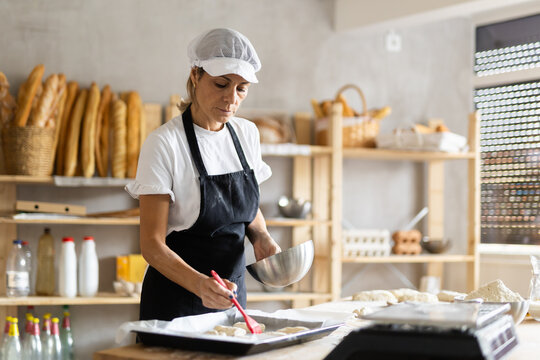 Woman brushing uncooked dough pieces on baking sheet with oil before baking in oven