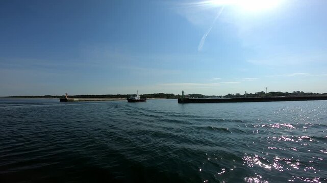 Motor vessel entering a harbour between breakwaters marked by red and green navigation beacons, with a trailing wake, rippling blue water and sunlight reflecting across the coastal port entrance