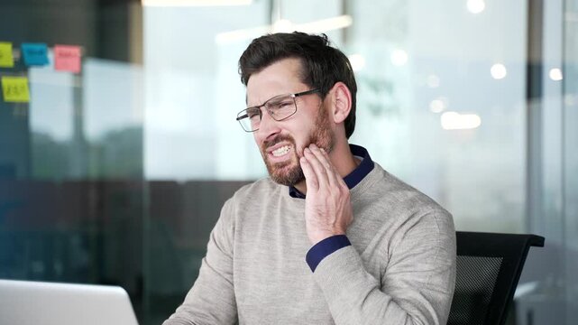 Close up of businessman suffering from toothache while sitting at desk in modern office. Mature professional holding hand on cheek, feeling sharp dental pain and discomfort, reacting to aching tooth.