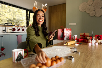 Smiling woman in reindeer antlers holding smartphone while selecting eggs for baking © Home-stock