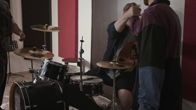 Tilt up shot of Caucasian young adult musicians entering rehearsal studio while drummer assembling cymbal on stand. Visual fits music academy campaigns and band promotion content