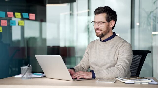 Pleasantly surprised professional smiles after reading good news on laptop. Happy mature businessman sitting at desk in modern office reacts to positive message on computer with visible satisfaction.